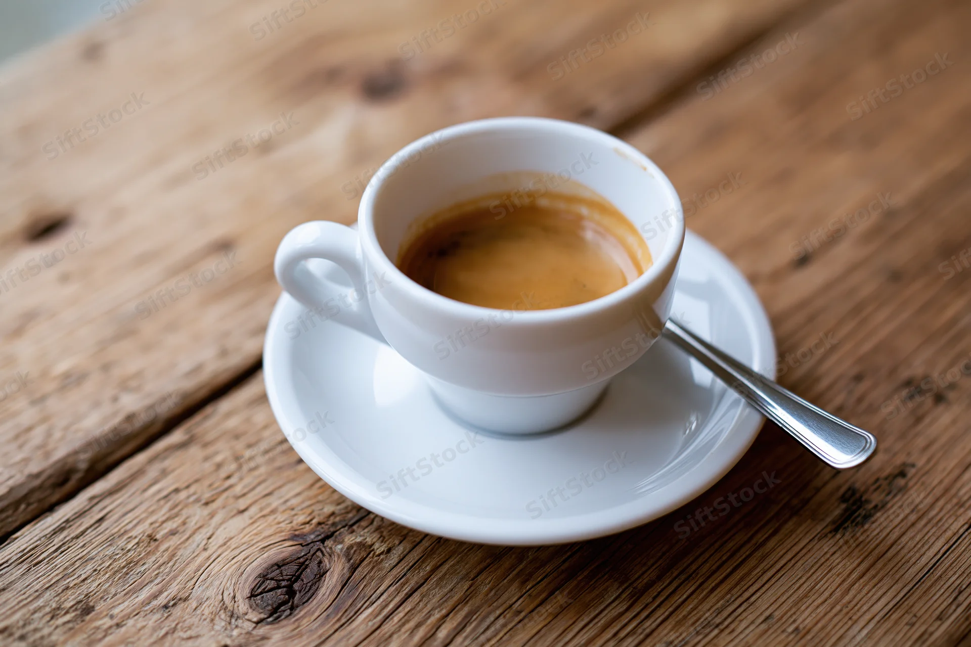 White ceramic espresso cup with tan foam on a wooden table, accompanied by a metal spoon and saucer