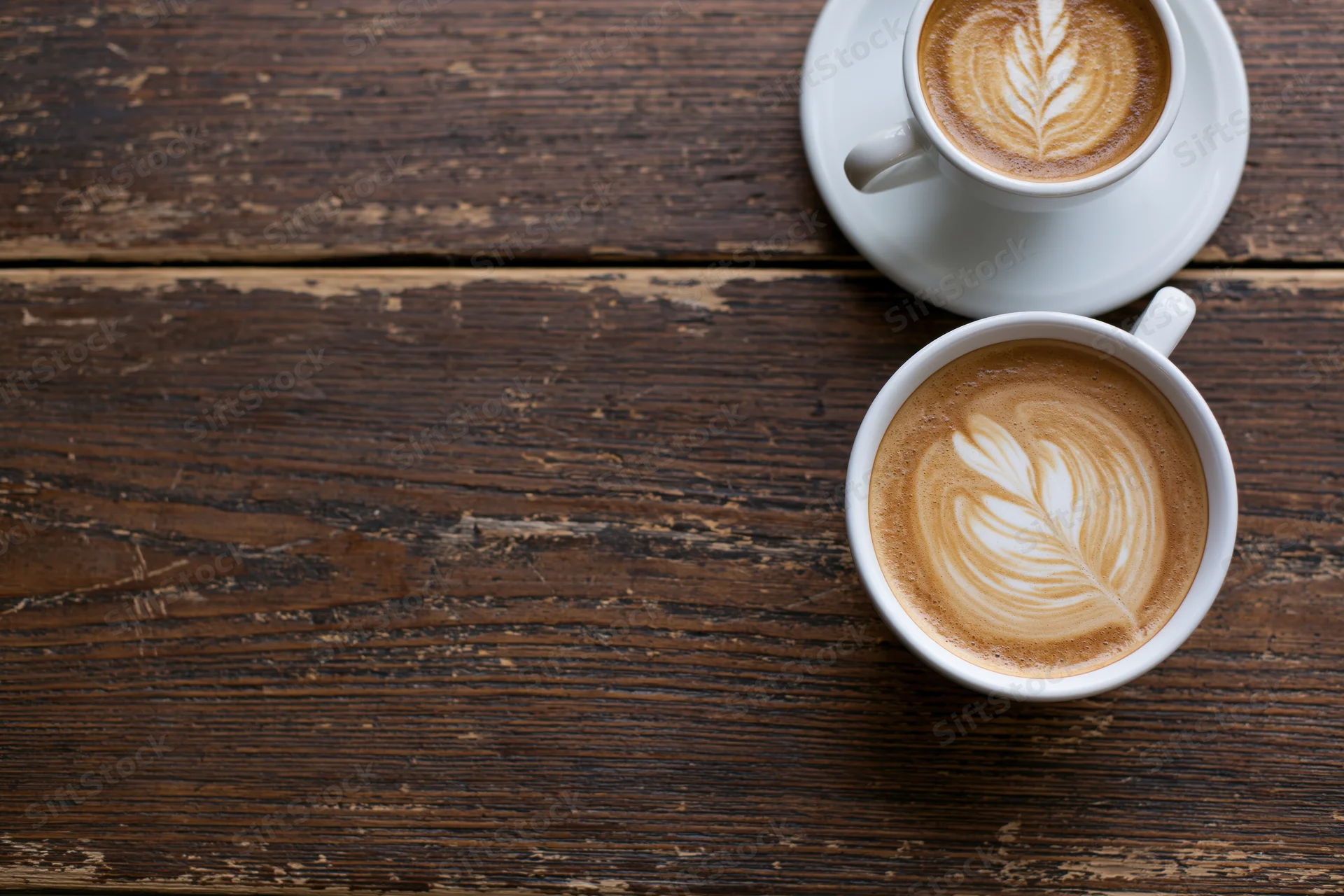 Two white ceramic cups of latte with leaf latte art on a rustic wooden table