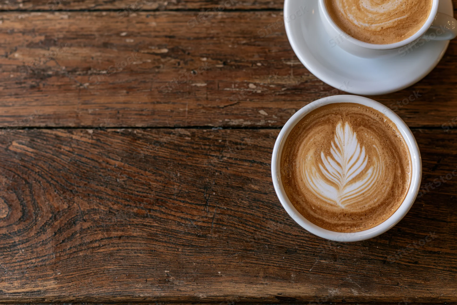 Two white ceramic cups of latte with leaf latte art on a rustic wooden table