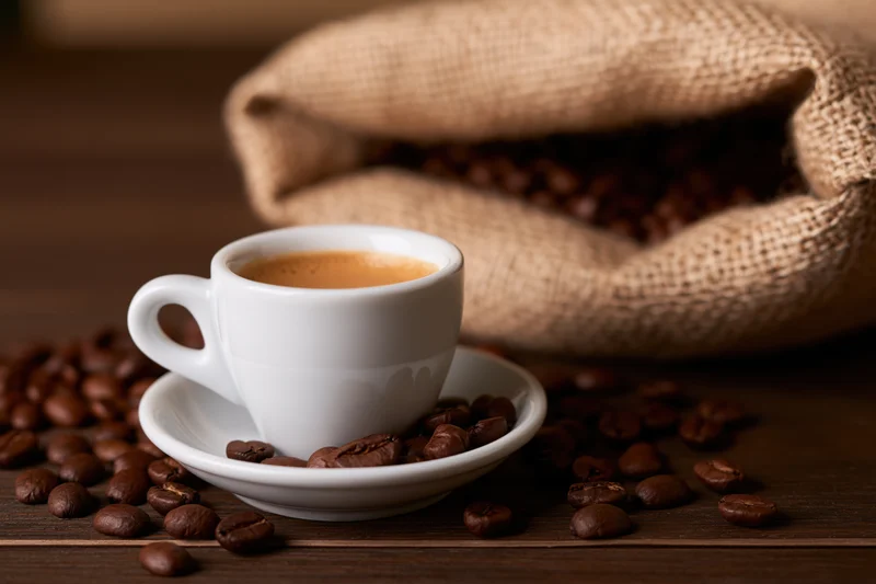 White ceramic espresso cup with crema on saucer surrounded by roasted coffee beans on dark wooden table near burlap sack