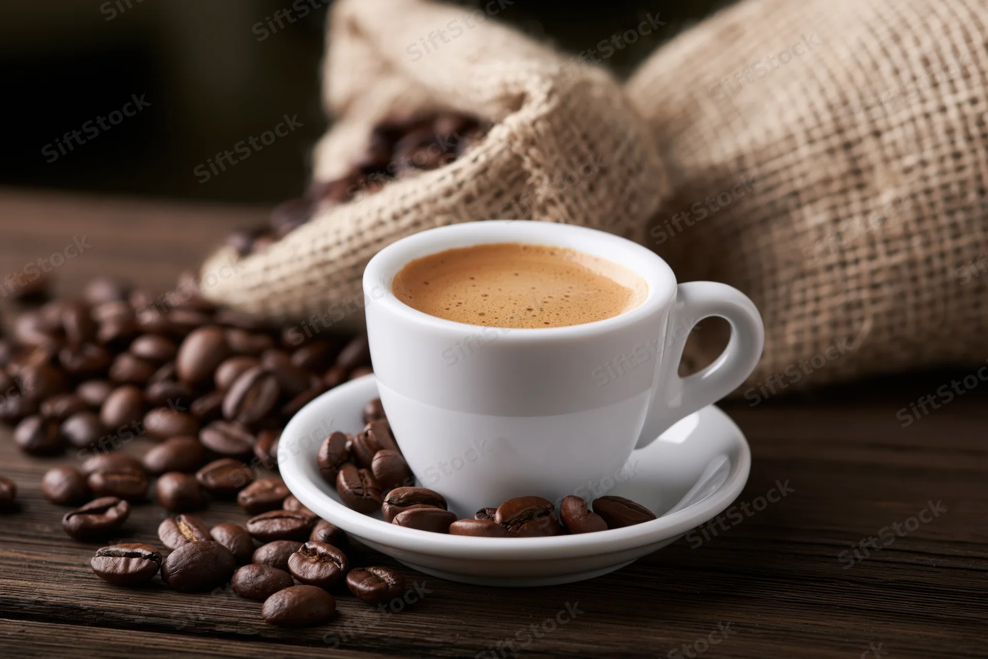 White ceramic espresso cup with crema on saucer surrounded by roasted coffee beans on dark wooden table near burlap sack