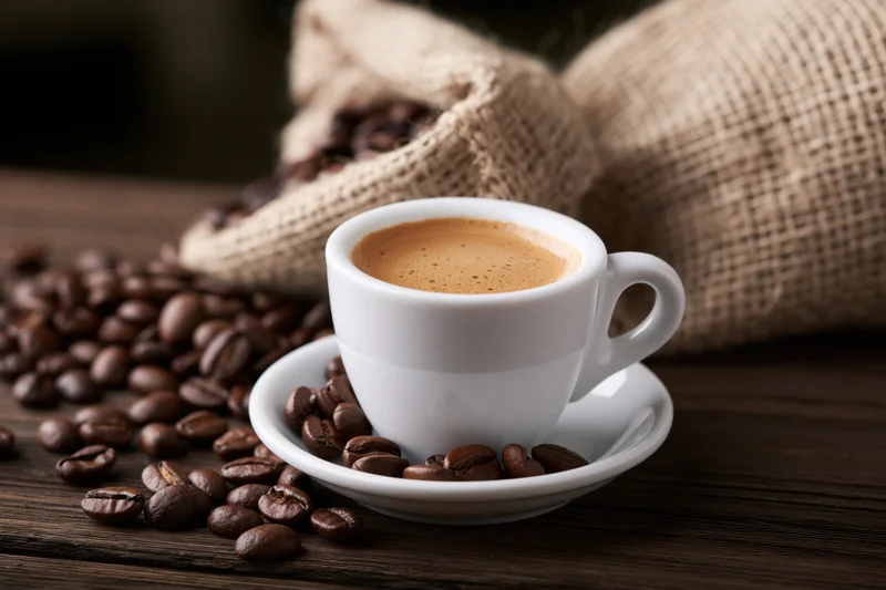 White ceramic espresso cup with crema on saucer surrounded by roasted coffee beans on dark wooden table near burlap sack