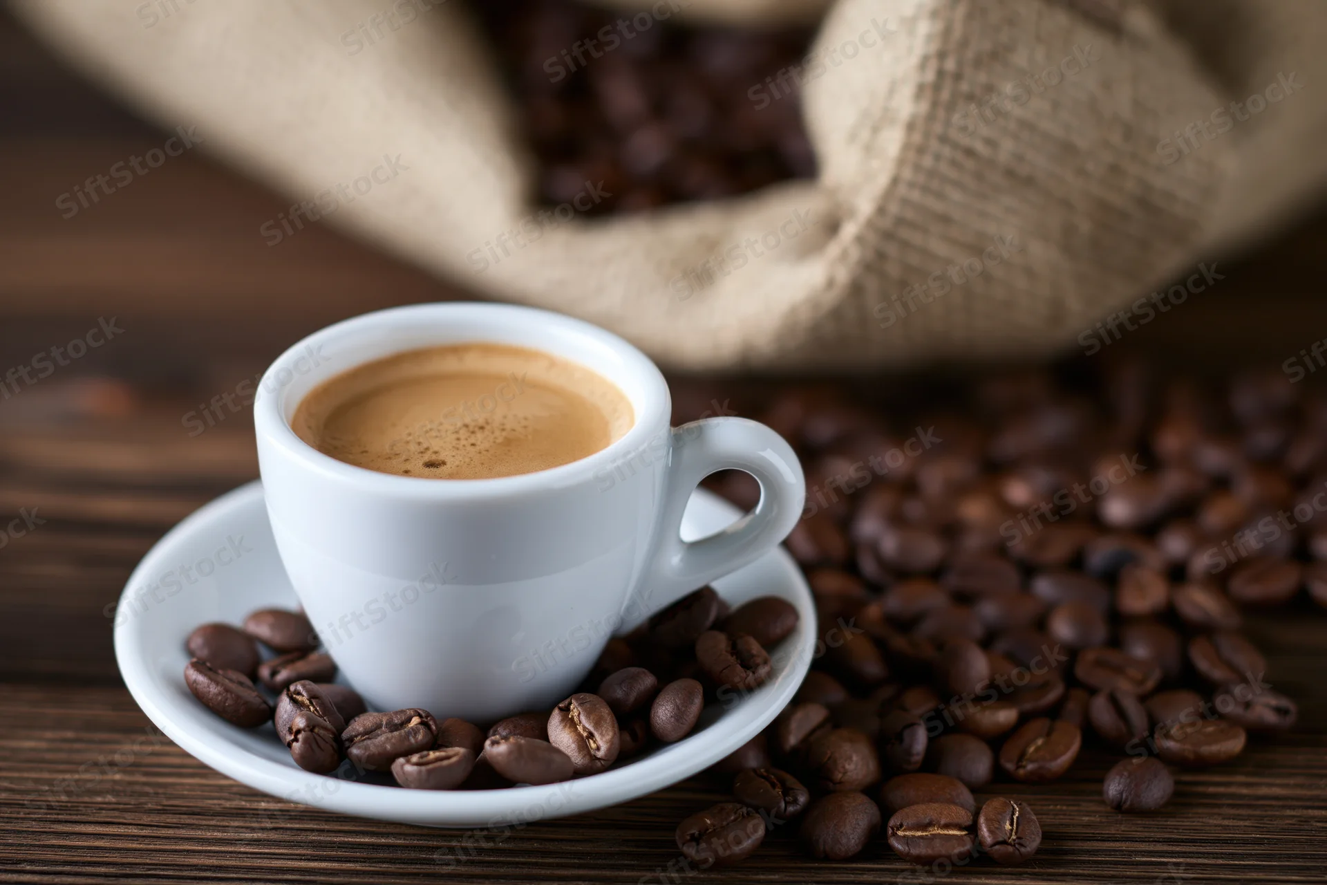 White ceramic espresso cup with crema on saucer surrounded by roasted coffee beans on wooden table near burlap sack