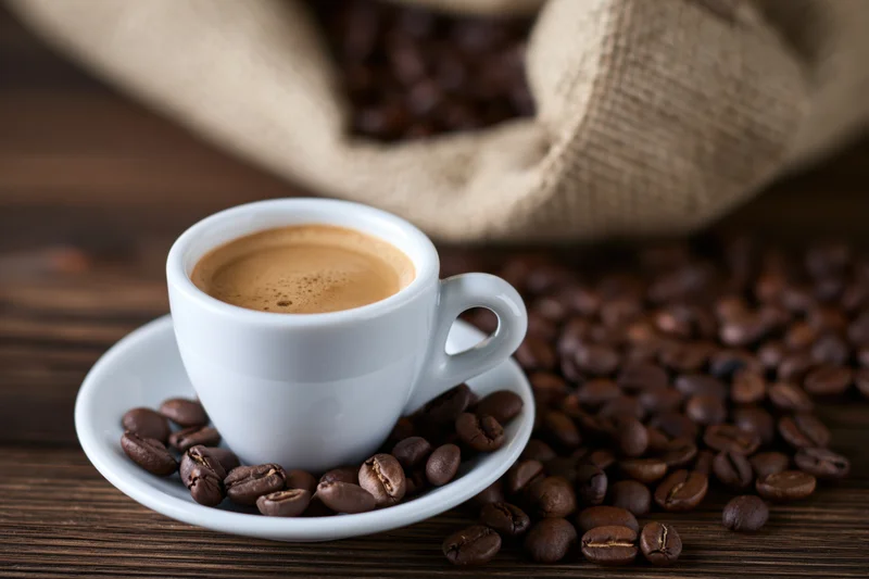 White ceramic espresso cup with crema on saucer surrounded by roasted coffee beans on wooden table near burlap sack