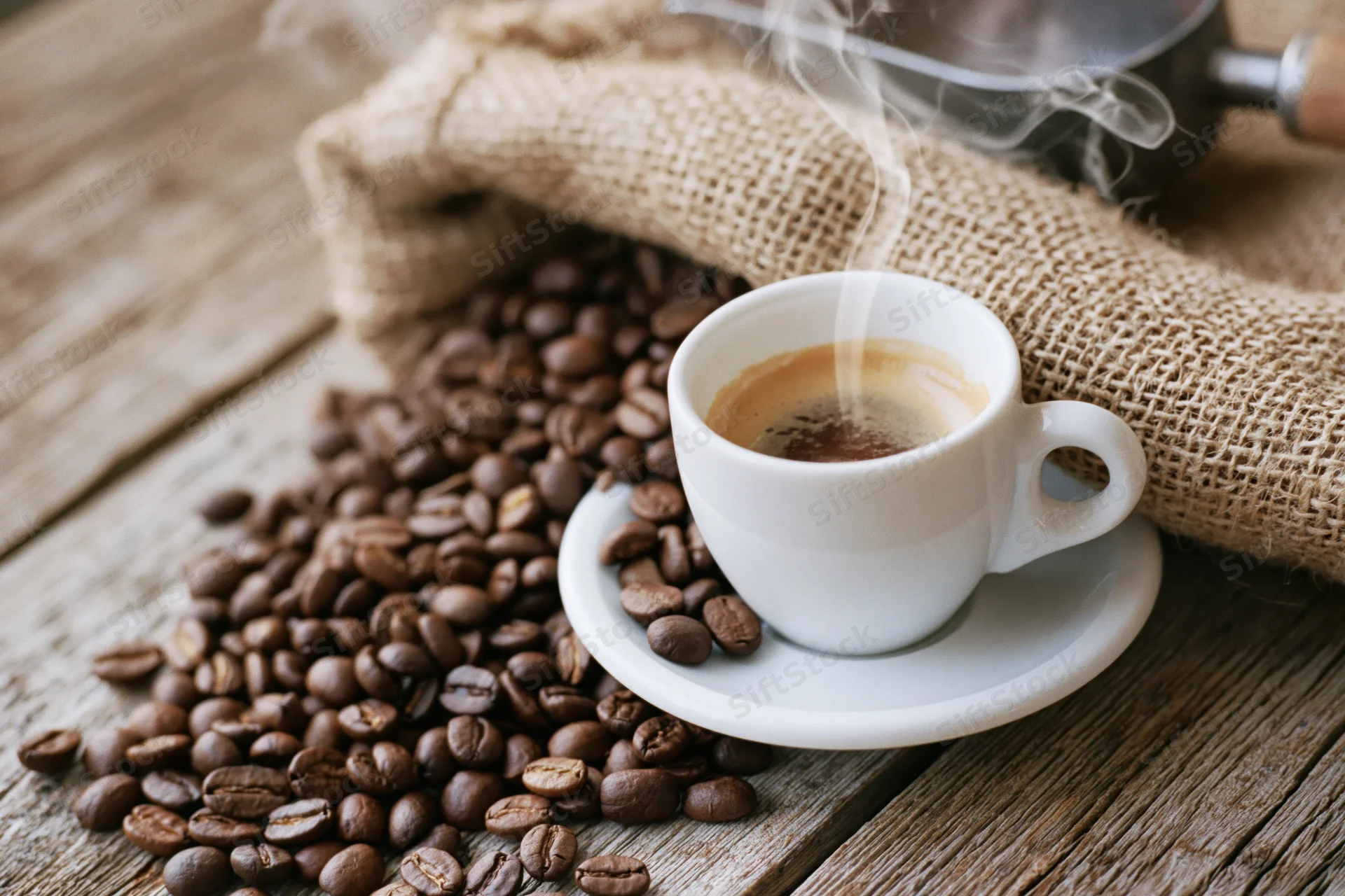 White ceramic espresso cup with steam on saucer surrounded by roasted coffee beans on wooden table