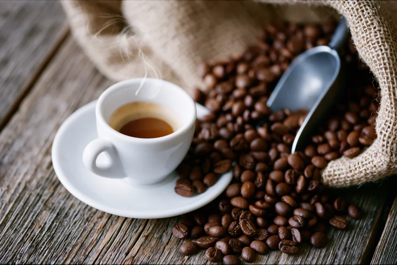 White ceramic espresso cup with steam on saucer beside roasted coffee beans spilling from burlap sack on wooden table