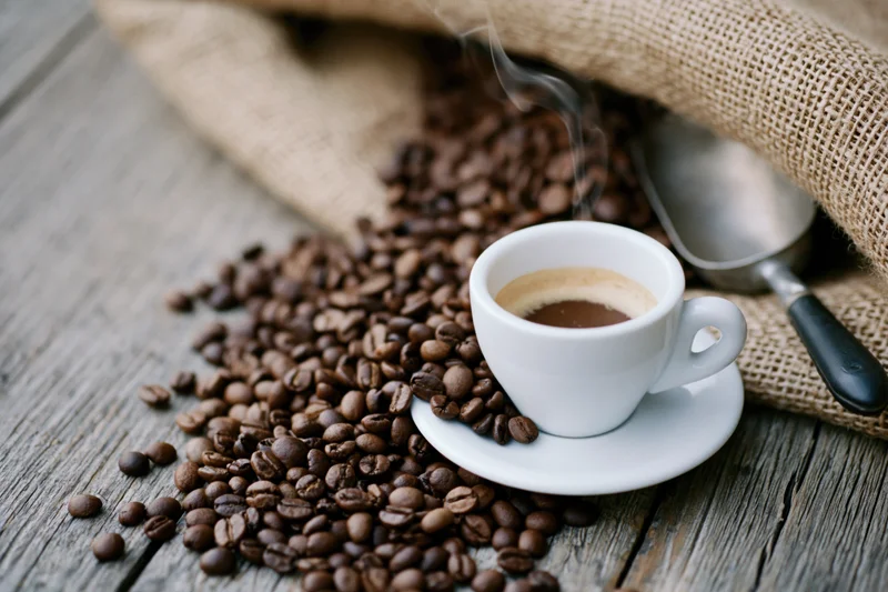 Steaming espresso cup on saucer surrounded by roasted coffee beans on wooden table with burlap sack and metal spoon