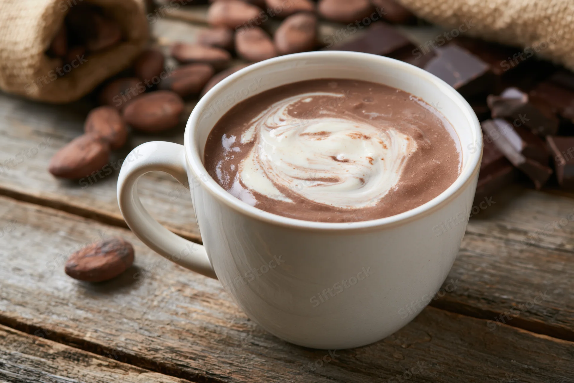 White ceramic mug filled with hot chocolate and cream swirl on wooden table surrounded by cocoa beans and chocolate chunks