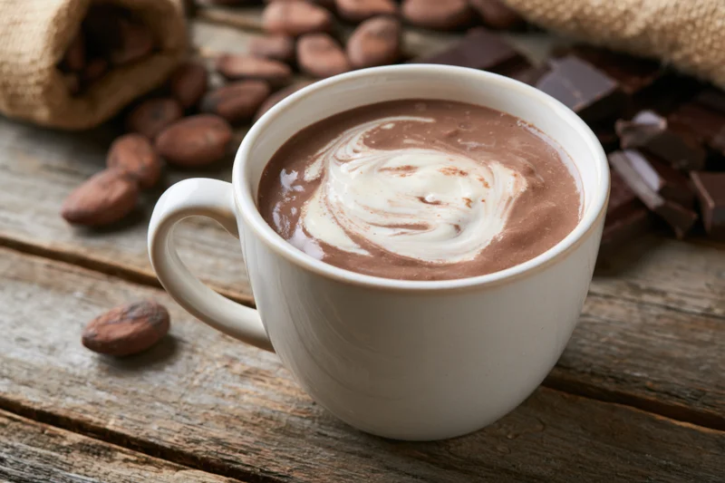 White ceramic mug filled with hot chocolate and cream swirl on wooden table surrounded by cocoa beans and chocolate chunks