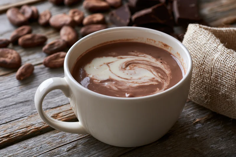 White ceramic mug filled with hot chocolate featuring cream swirl on wooden table with cocoa beans and burlap sack