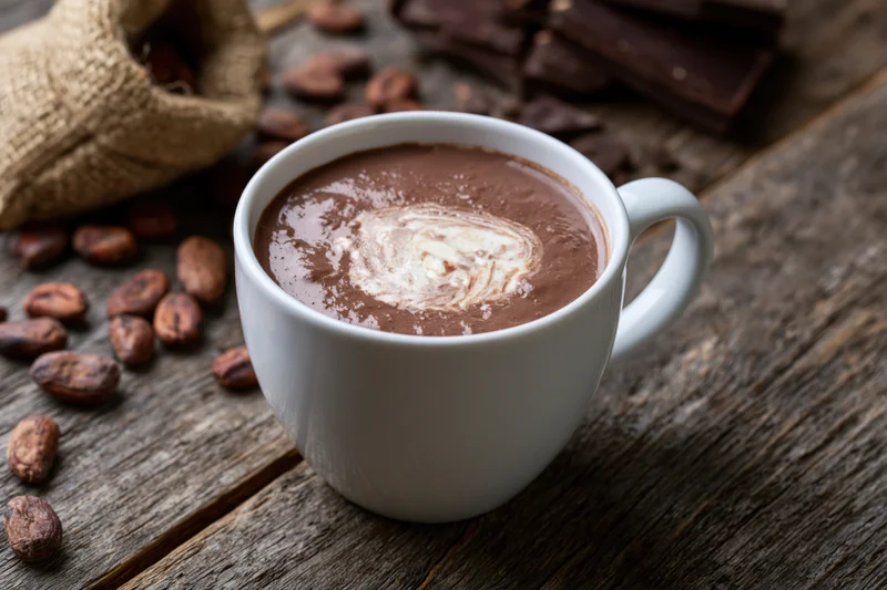 White ceramic mug filled with hot chocolate featuring cream swirl on rustic wooden table surrounded by cocoa beans and chocolate bars