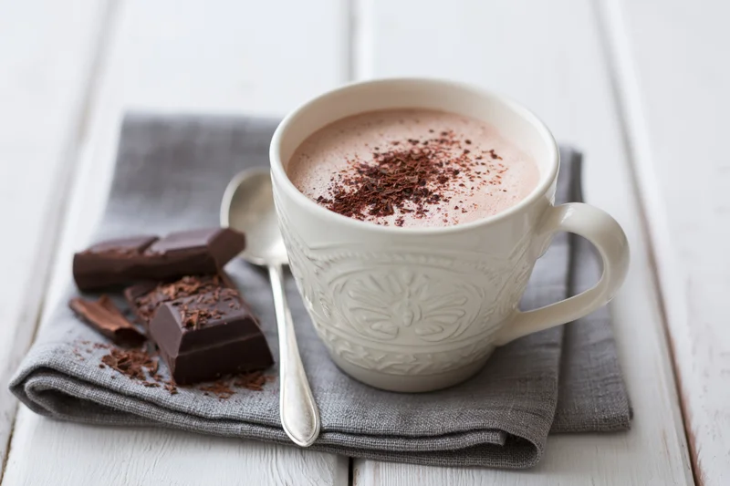 White embossed ceramic mug filled with frothy hot chocolate topped with cocoa shavings on a gray napkin with chocolate pieces and spoon beside it on a white wooden table