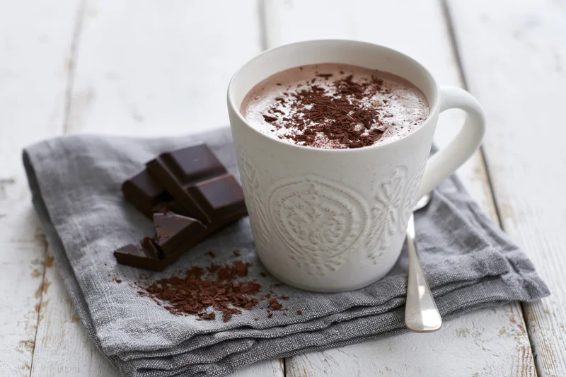 White embossed ceramic mug filled with frothy hot chocolate on a gray napkin with chocolate pieces and spoon on a wooden table