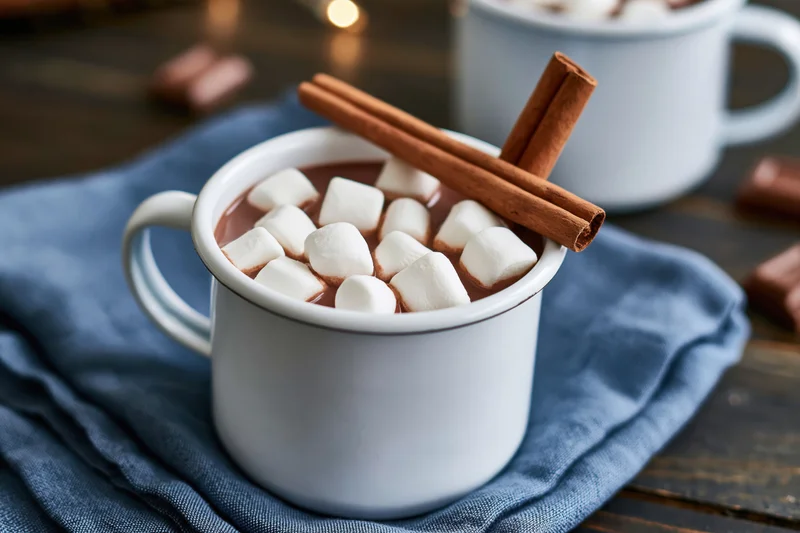 White enamel mug filled with hot chocolate topped with marshmallows and cinnamon sticks on a blue cloth over wooden table