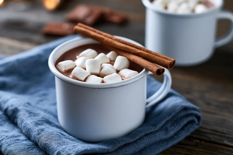 White enamel mug filled with hot chocolate topped with marshmallows and cinnamon sticks on a blue cloth over wooden table