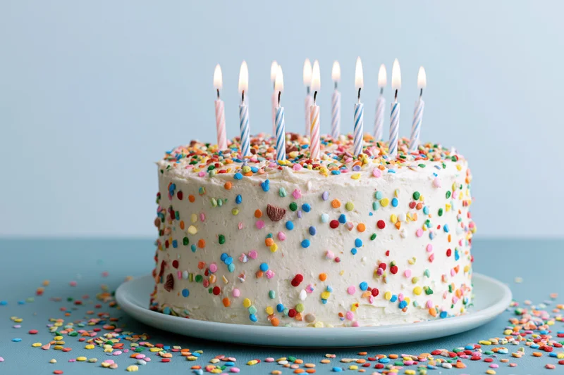White frosted birthday cake with lit candles and colorful sprinkles on a blue surface