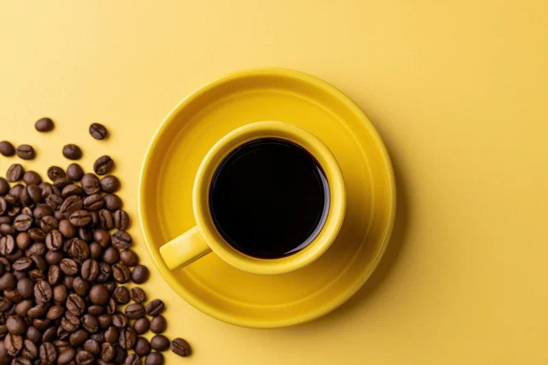 Yellow ceramic coffee cup filled with black coffee on saucer beside scattered coffee beans on yellow surface