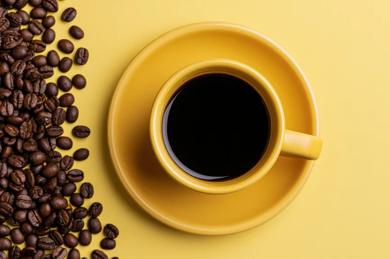 Yellow ceramic coffee cup filled with black coffee on saucer beside scattered coffee beans on yellow surface