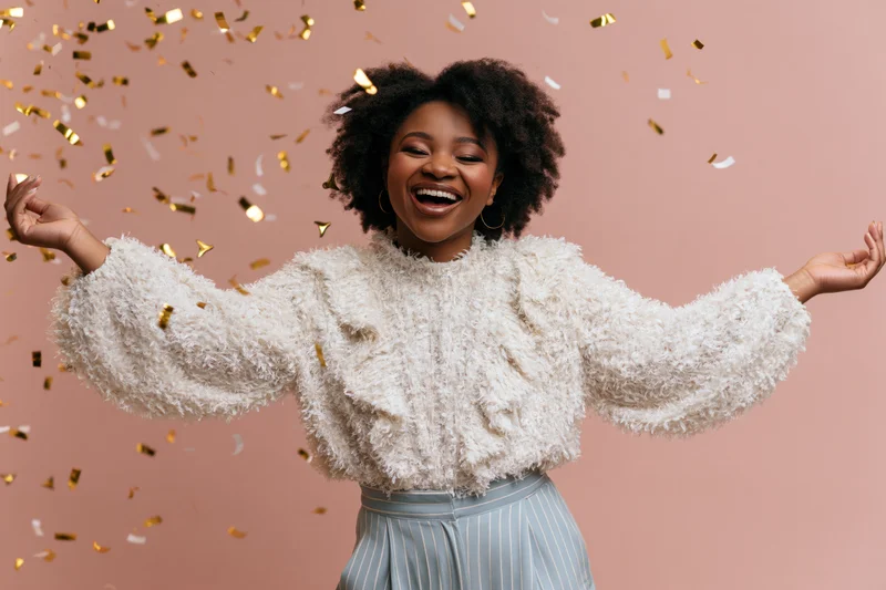 Young black woman laughing with arms spread amidst falling confetti against pink background