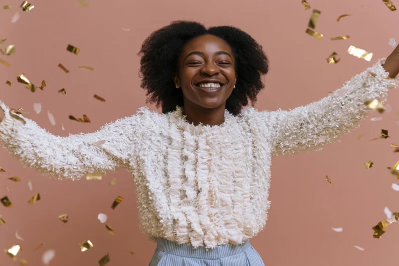 Young black woman laughing with arms spread amidst falling gold confetti in studio setting