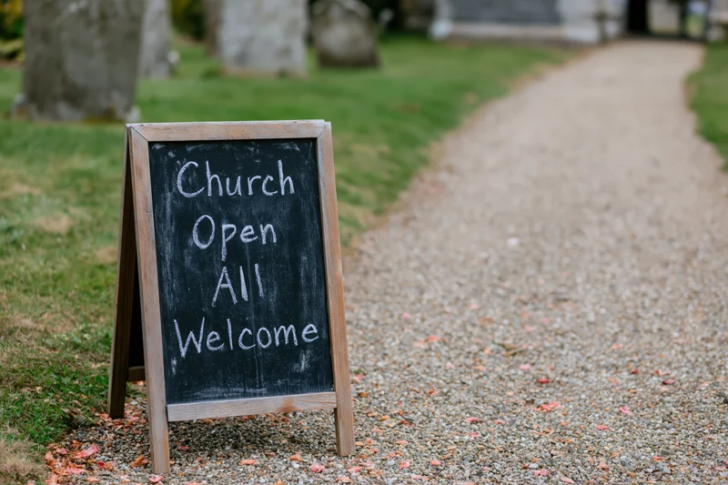 Aframe chalkboard sign reading church open all welcome on gravel path near church entrance