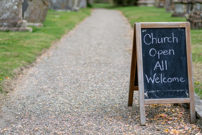 Aframe chalkboard sign reading church open all welcome on gravel path beside cemetery grass