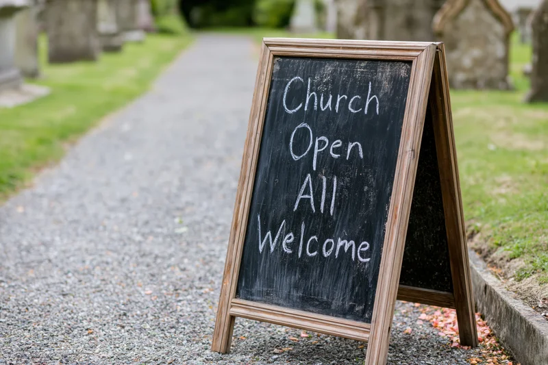 Aframe chalkboard sign reading church open all welcome on gravel path in cemetery