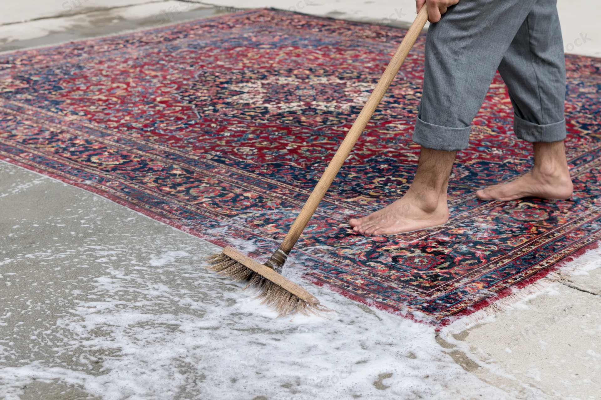 Barefoot man scrubbing a large wet Persian rug laid flat on concrete with a wooden broom and soap foam