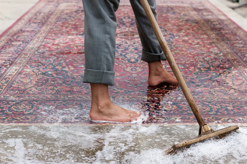 Barefoot man scrubbing a large wet Persian rug laid flat on pavement with a wooden broom and soapy water