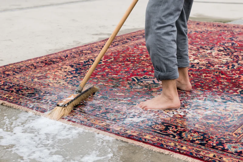Barefoot man scrubbing a large wet persian rug laid flat on concrete pavement