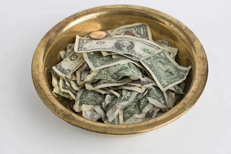 Crumpled us dollar bills and coins in a brass offering plate on white background