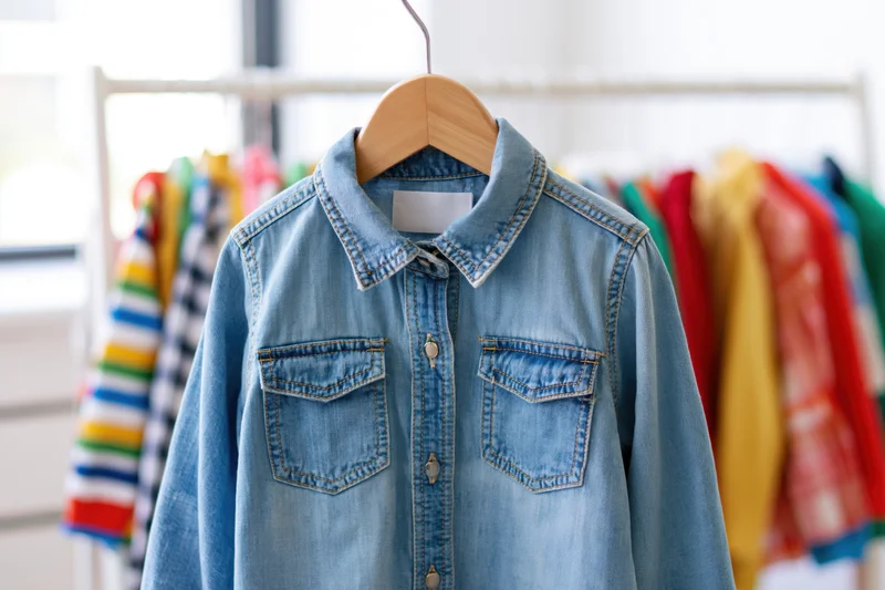 Childrens denim button-up shirt hanging on wooden hanger in front of clothing rack