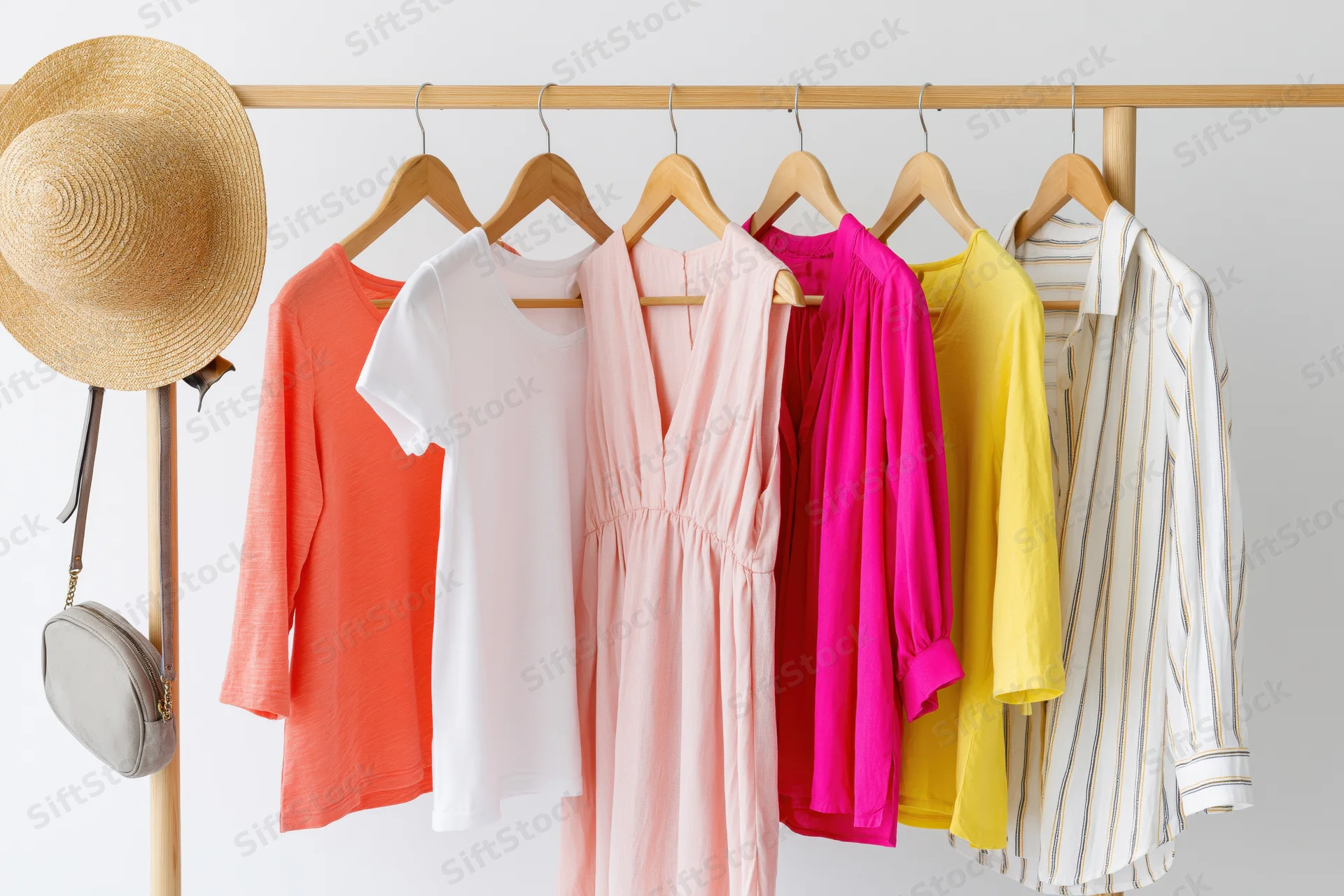 Six women's summer garments including dresses and tops hanging on a wooden rack with a straw hat and bag attached