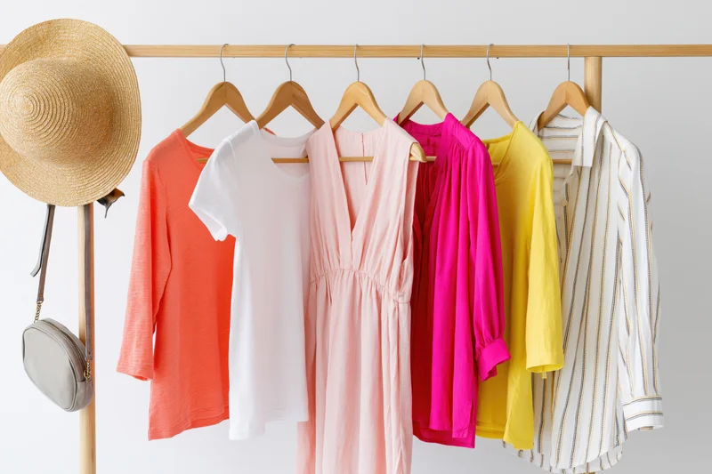 Six women's summer garments including dresses and tops hanging on a wooden rack with a straw hat and bag attached