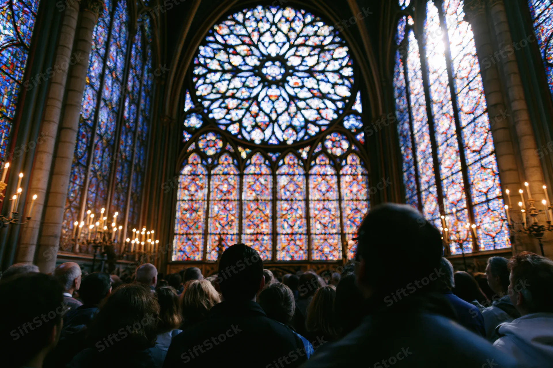 Visitors in silhouette facing a large stained glass rose window inside a gothic cathedral