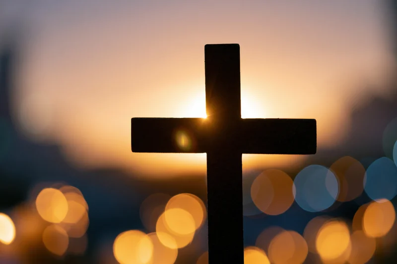Dark silhouetted christian cross against a glowing sunset sky with bokeh lights