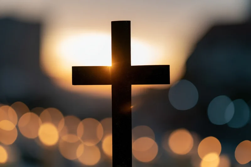 Silhouetted christian cross against a glowing sunset sky with blurred city lights below