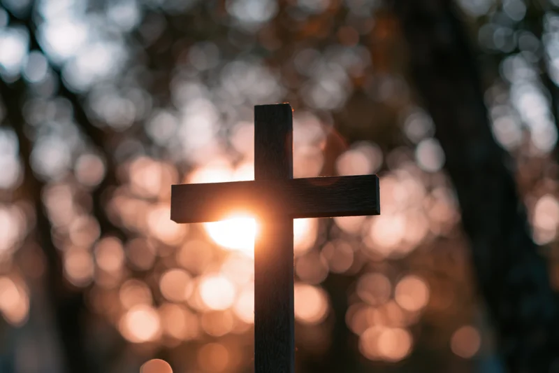 Dark wooden christian cross silhouetted against warm sunset light in forest setting