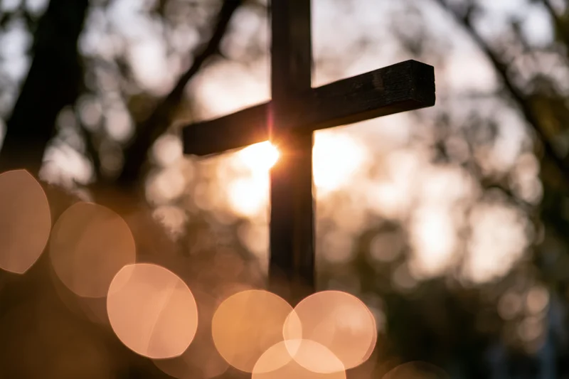 Wooden christian cross silhouetted against a glowing sunset with bokeh lights in a forest setting