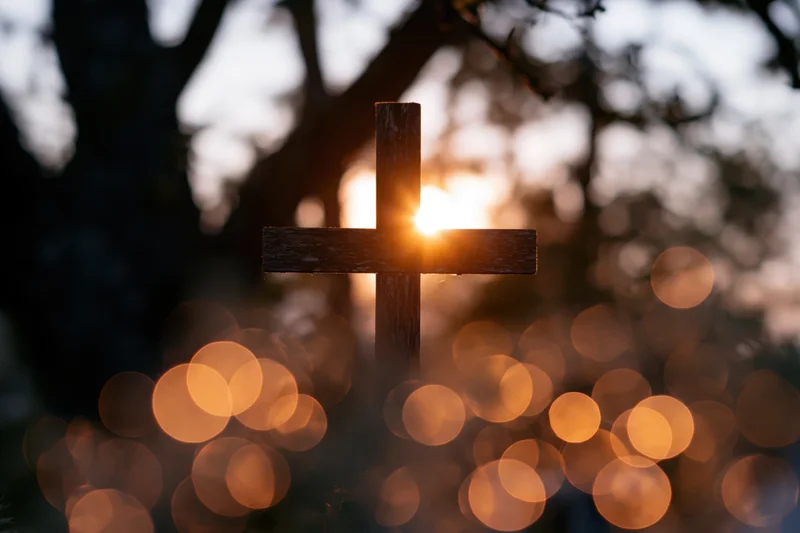 Wooden christian cross silhouetted against sunset with bokeh lights in background