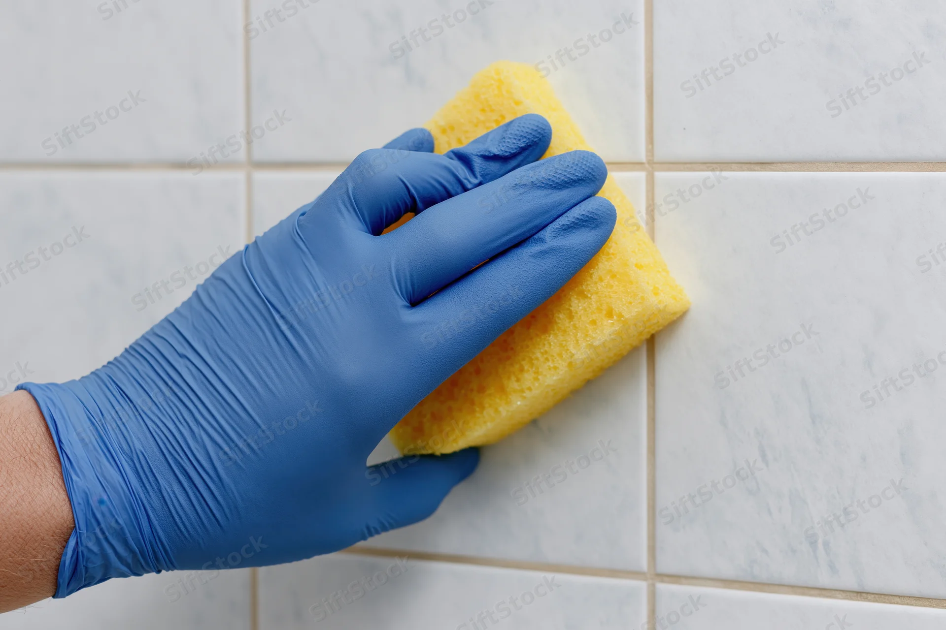 Hand in blue rubber glove scrubbing white tiled wall with yellow sponge