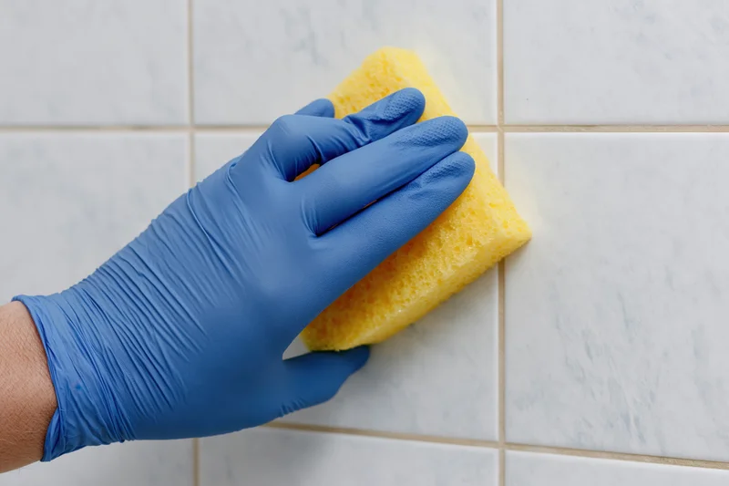 Hand in blue rubber glove scrubbing white tiled wall with yellow sponge