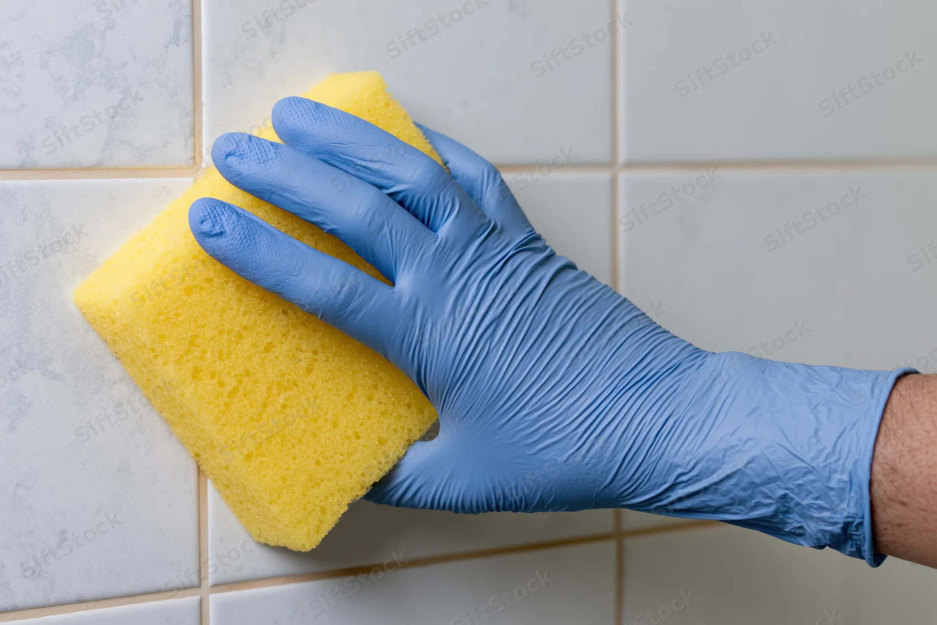 Hand in blue rubber glove scrubbing white tiled surface with yellow sponge