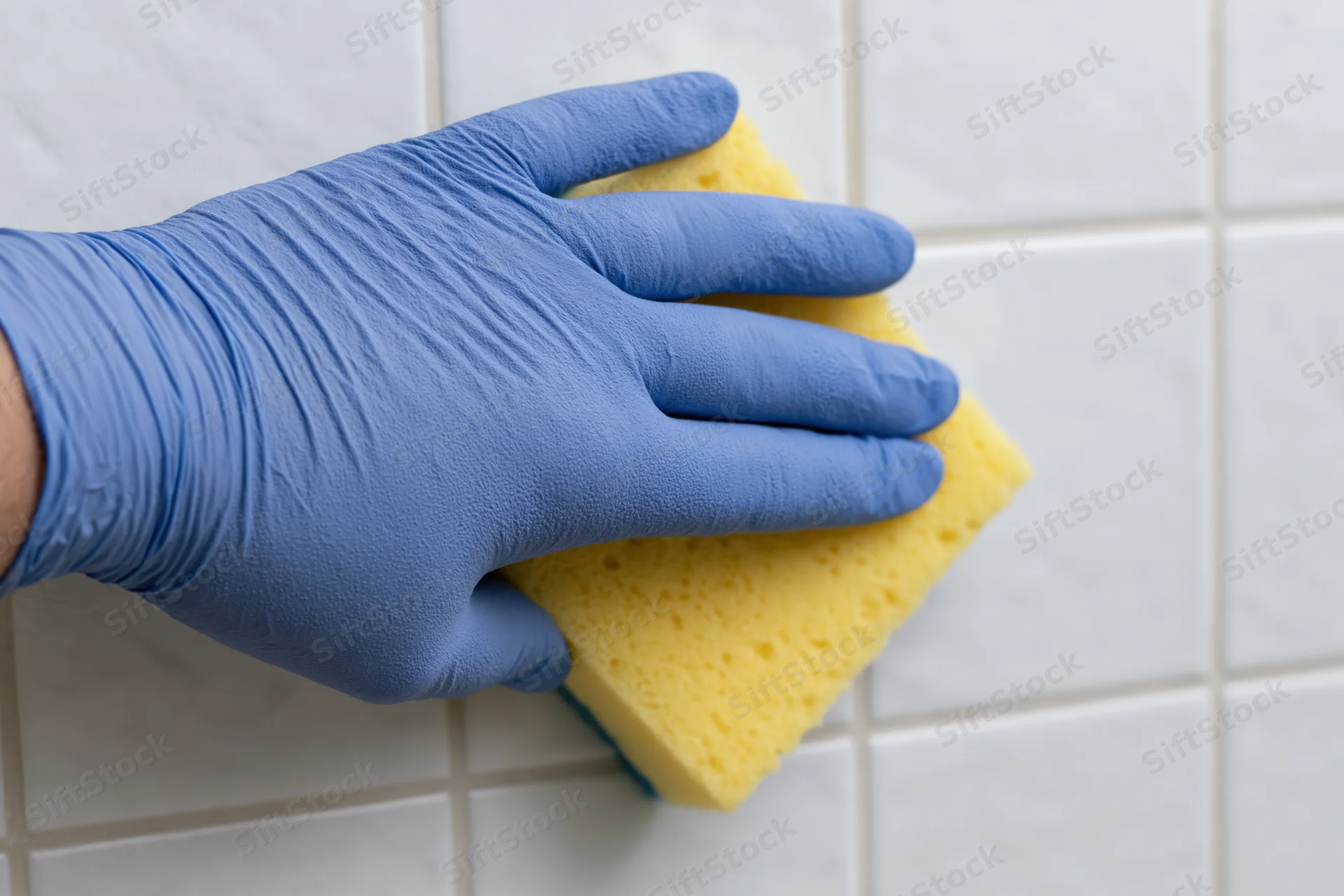 Hand in blue latex glove scrubbing white tile with yellow sponge