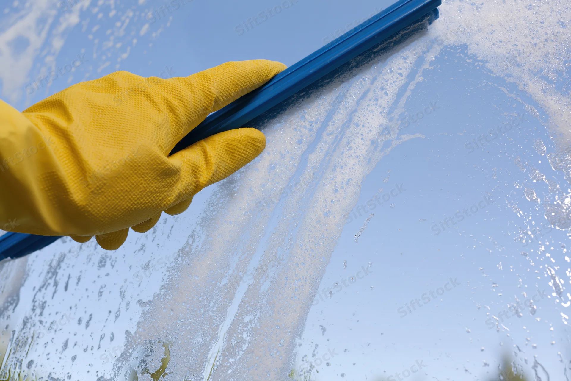 Hand in yellow rubber glove using blue squeegee to clean soapy window outdoors