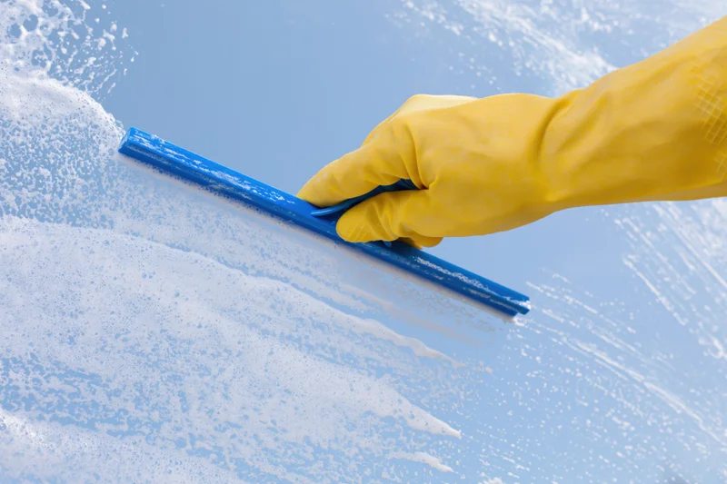 Hand in yellow rubber glove using blue squeegee to clean soapy window glass outdoors