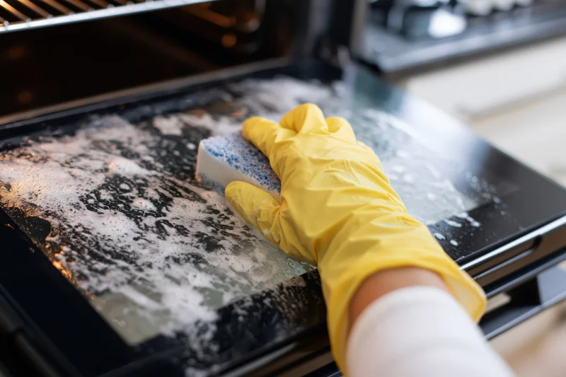 Hand in yellow glove scrubbing soapy oven door with sponge in kitchen