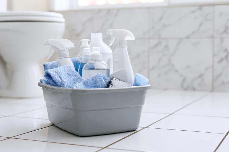 Gray plastic basin filled with cleaning supplies and blue towels on tiled bathroom floor