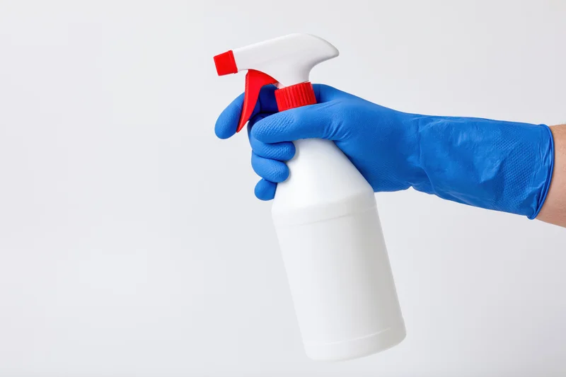 Hand in blue rubber glove holding white spray bottle against plain background