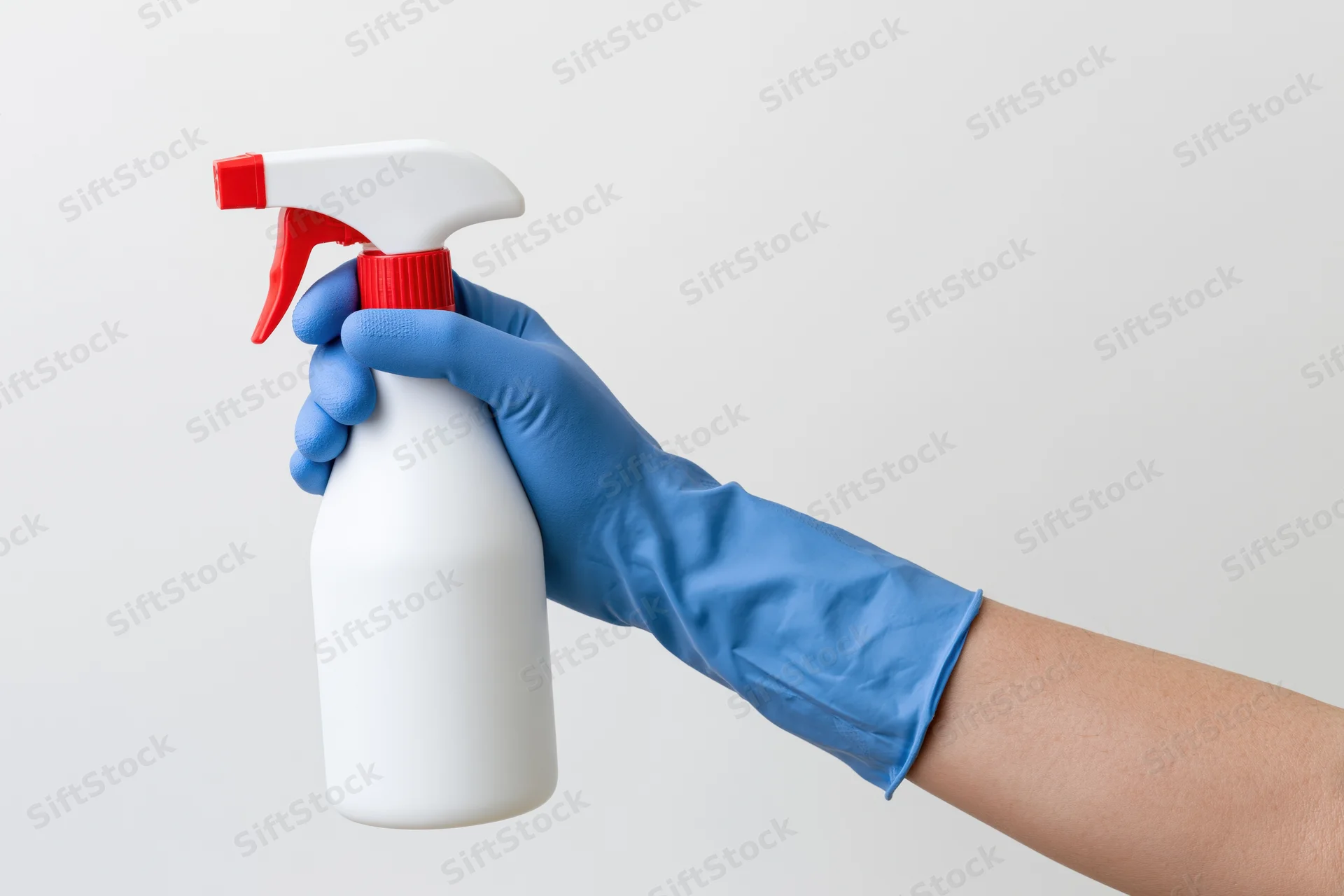 Hand in blue cleaning glove holding white spray bottle with red nozzle against plain background